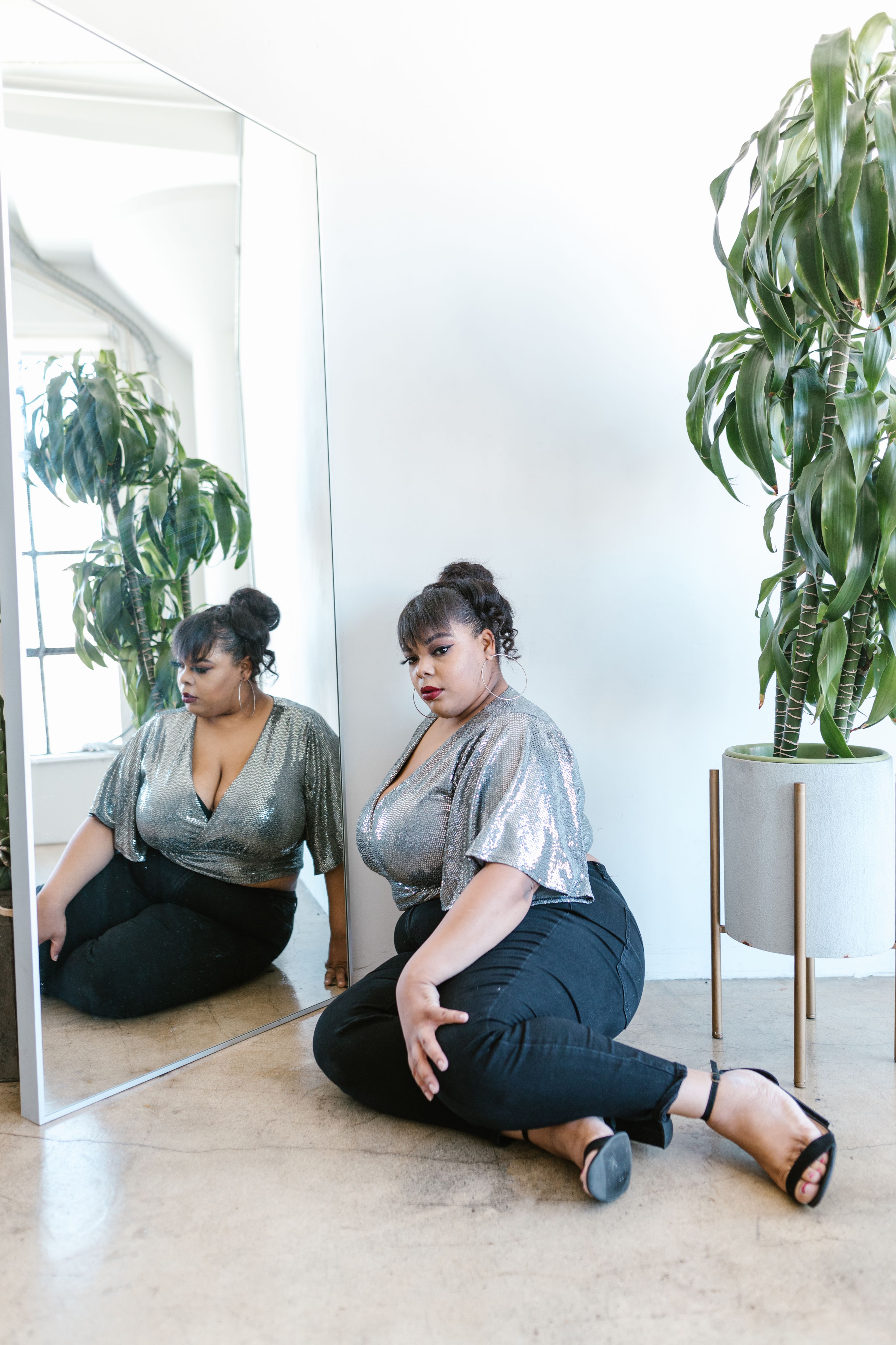 Mulher plus size estilosa em uma pose reflexiva ao lado de um grande espelho dentro de casa.