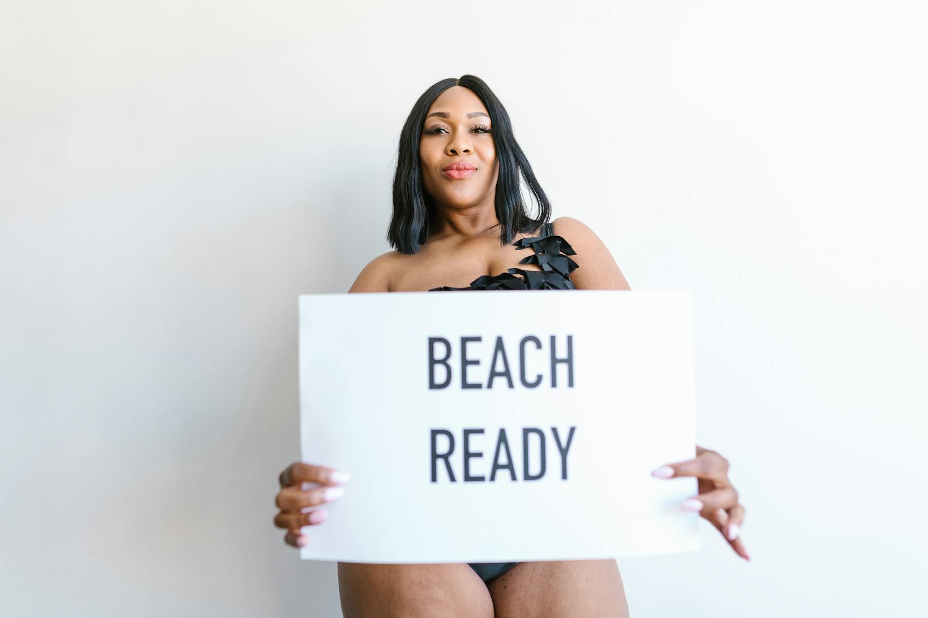 A confident woman in swimwear holding a 'BEACH READY' sign, embracing body positivity.