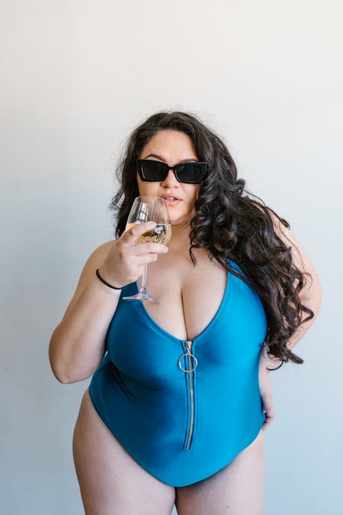 Woman in Blue Swimwear Holding a Glass Wine while Wearing Black Sunglasses on White Background
