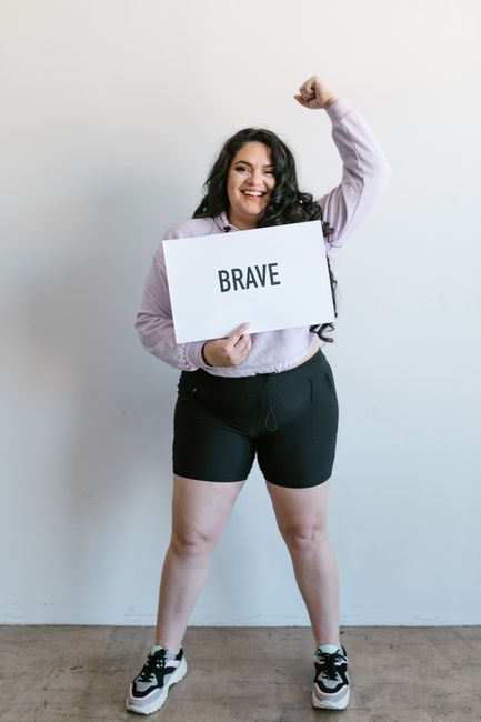 Curvy woman in casual attire holding a 'BRAVE' placard with a smile in a bright indoor setting.