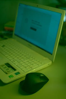 A white laptop with a black mouse on a desk, cast in a green tint, creating a digital workspace ambiance.