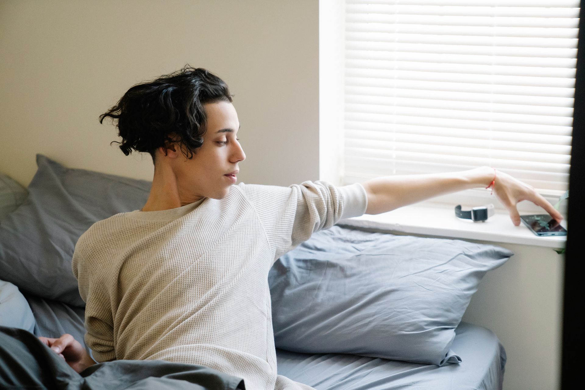 Young ethnic male adolescent taking mobile phone from windowsill after awakening in morning in bedroom