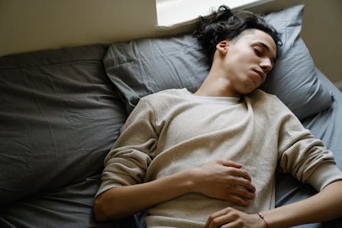 Free From above of relaxed millennial ethnic male in comfy clothes napping peacefully on soft bed with gray bedclothes Stock Photo