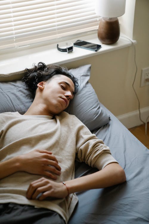 Free From above of young male in sleepwear sleeping on bed in bedroom with smartphone and wristwatch placed on windowsill in weekend morning Stock Photo