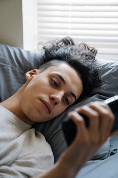 A young man relaxes on his bed, browsing his smartphone at home.