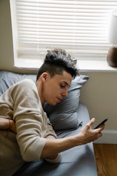 Young man lying in bed using a smartphone for online browsing in a cozy bedroom.