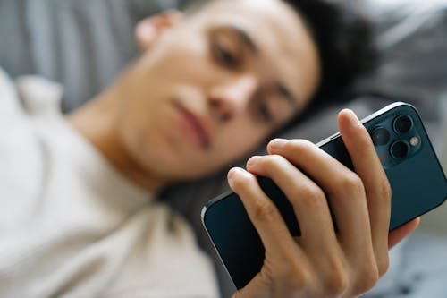 Soft focus of young guy lying on bed and using modern smartphone in weekend morning at home