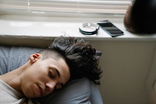 High angle of young Hispanic male with curly hair lying on bed near windowsill and sleeping in morning at home