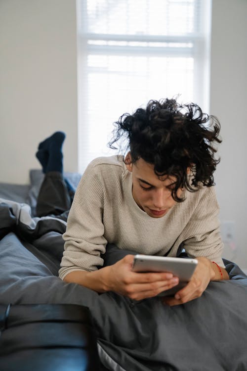 Free Young Hispanic man with curly hair lying on bed and browsing social media on tablet in weekend at home Stock Photo