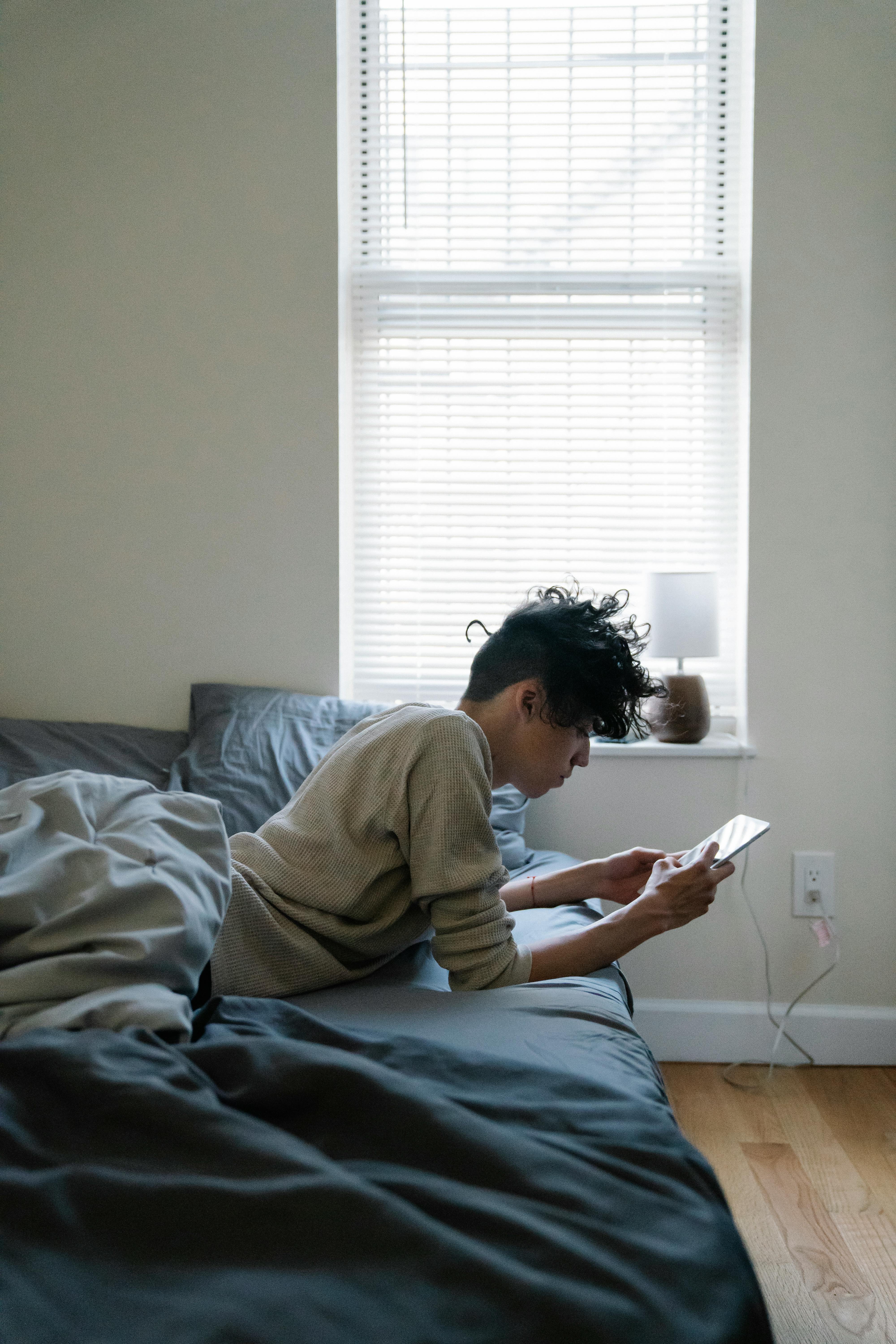 Young man lying on bed using smartphone in a modern indoor setting.