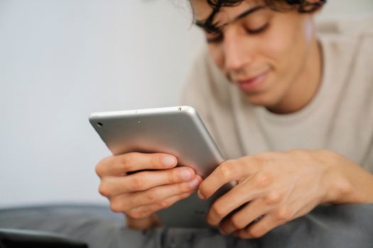 Crop happy ethnic male teenager in casual clothes smiling while lying on bed and using tablet during weekend at home