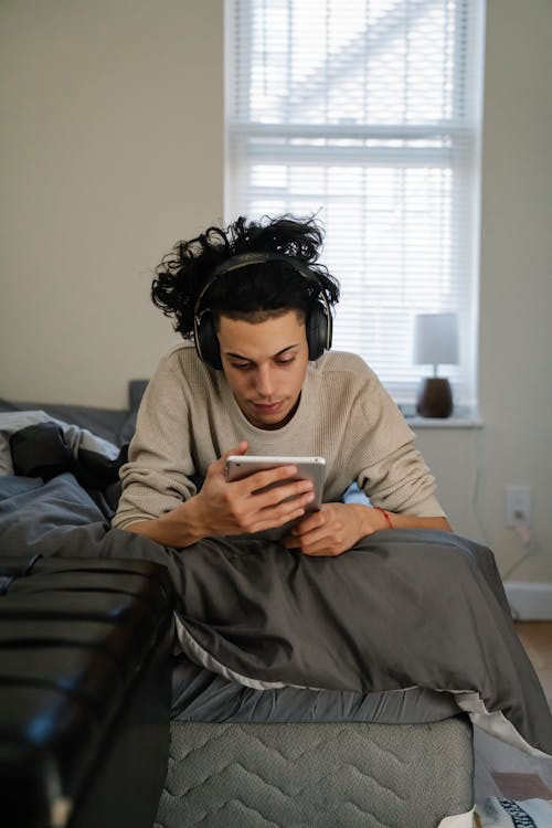 Free Young ethnic guy with dark hair in casual clothes listening to music and browsing tablet while sitting on bed at home Stock Photo