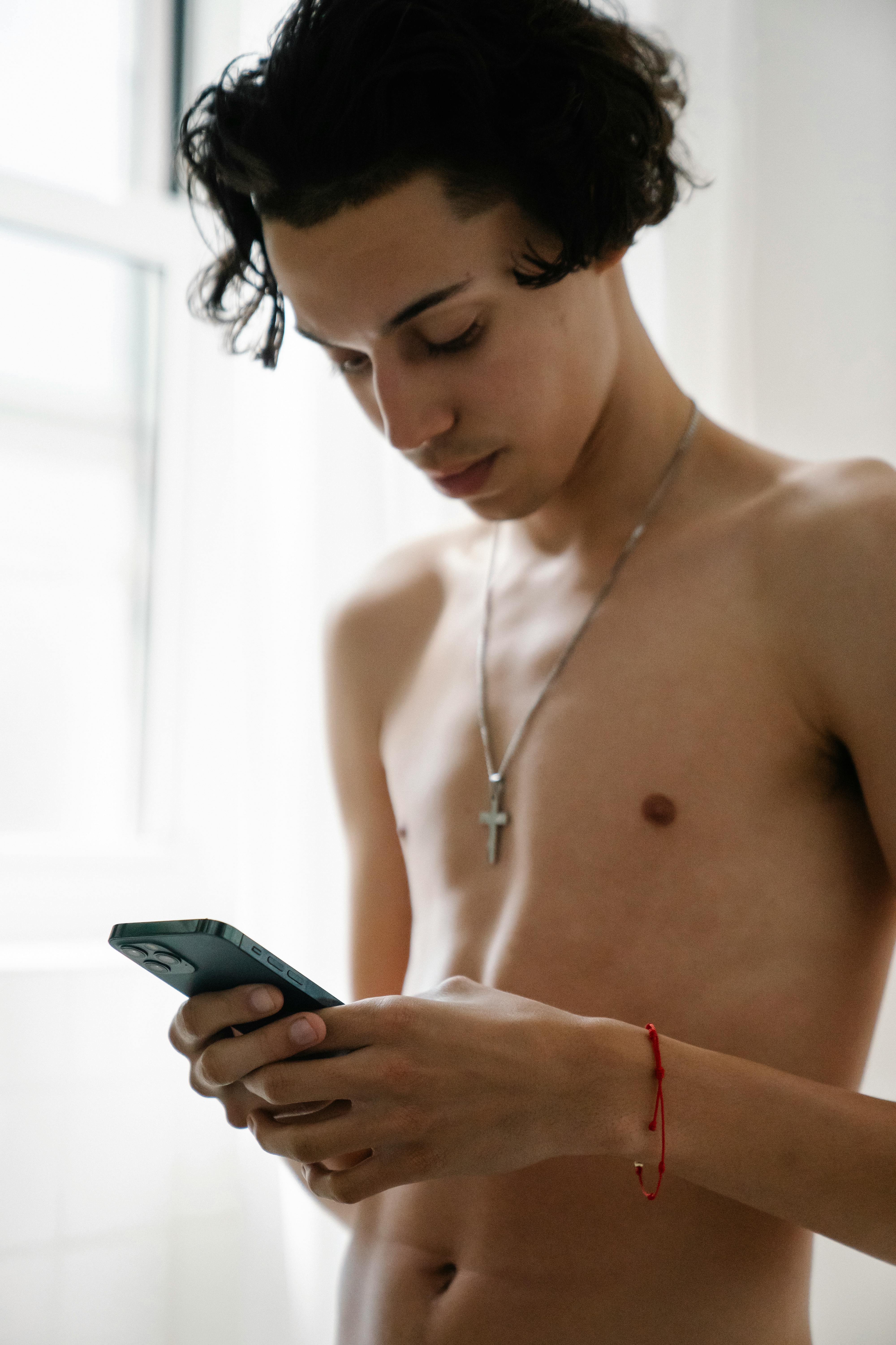 A shirtless young man uses his smartphone, standing in natural light with a cross necklace visible.