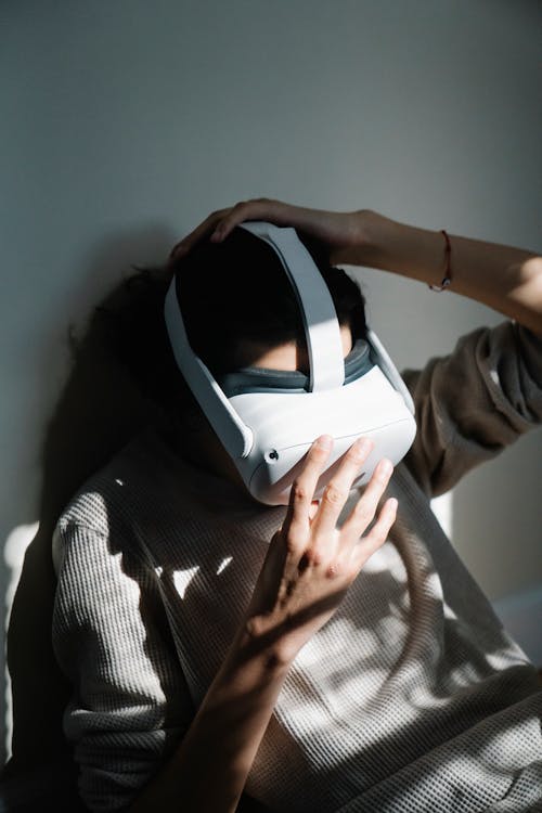 From above of unrecognizable male in casual clothes leaning on wall and adjusting modern headset while sitting on bed and exploring virtual reality