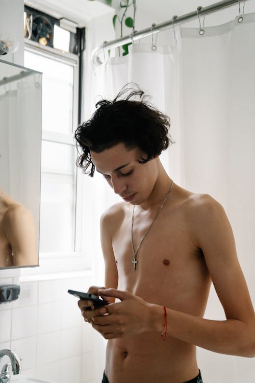 Young shirtless ethnic male messaging on mobile phone while standing in bathroom during morning hygienic routine