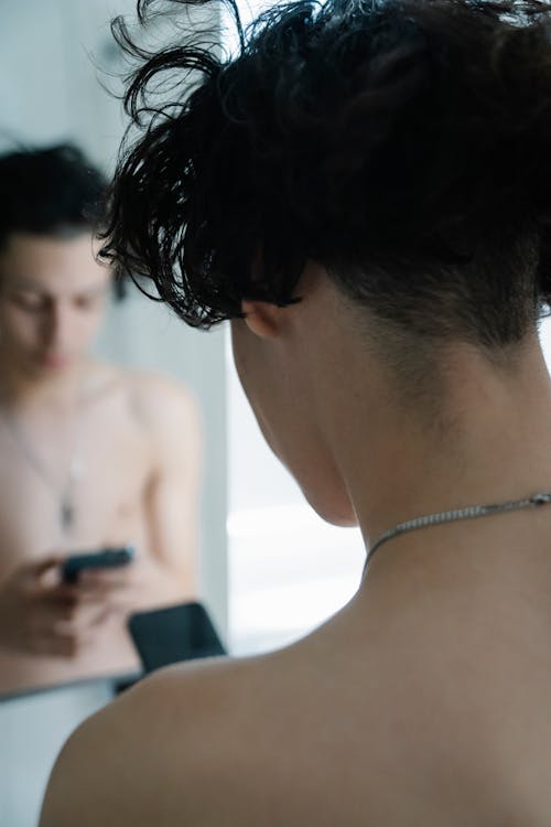 Young shirtless guy browsing smartphone near mirror in bathroom