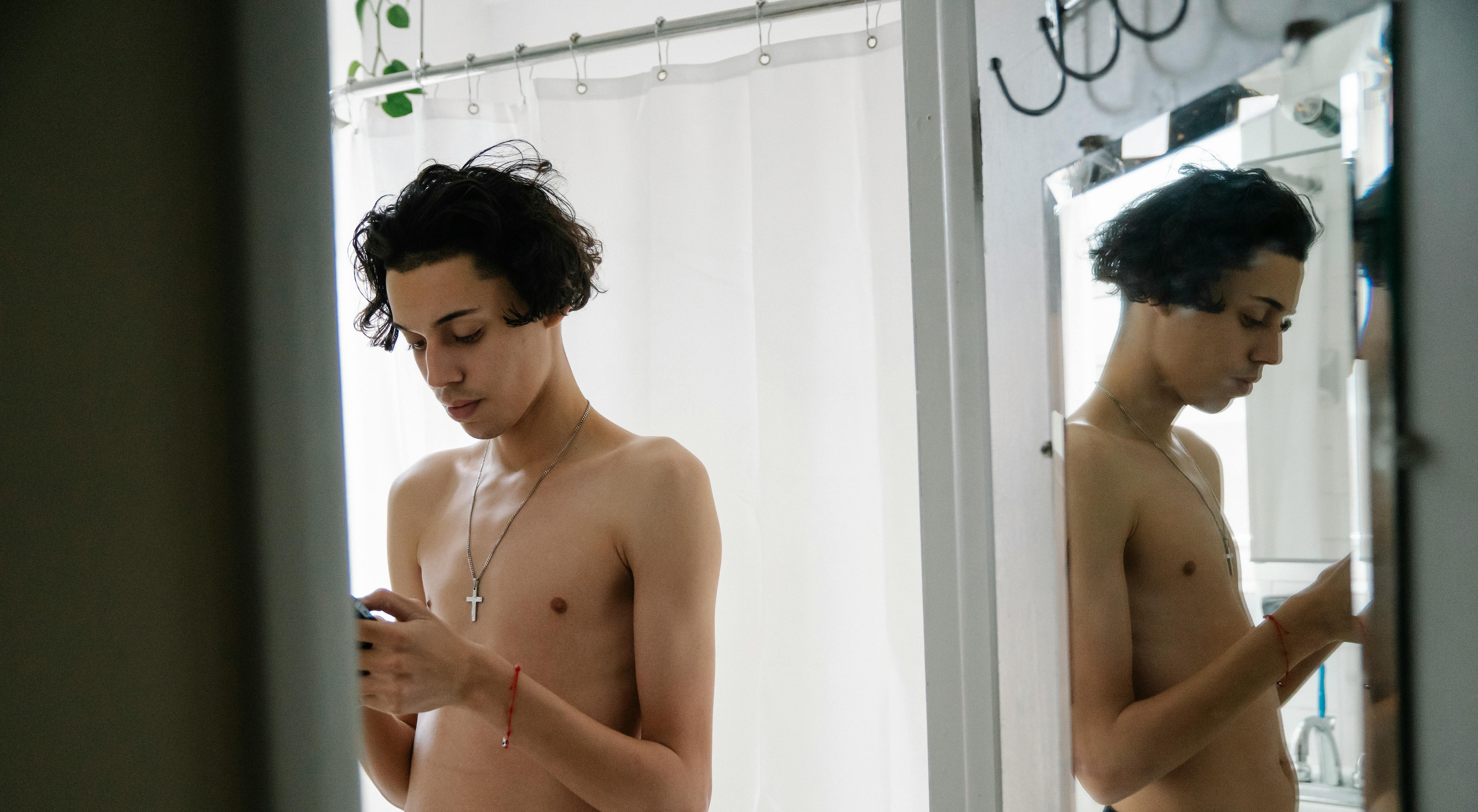 focused young ethnic male with naked torso messaging on smartphone in bathroom