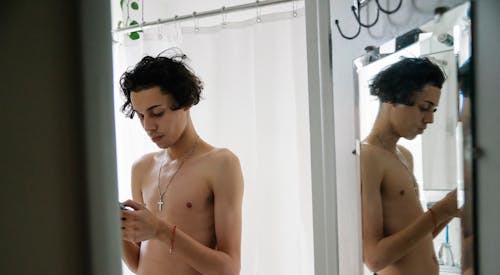 Concentrated young shirtless ethnic guy with dark hair using mobile phone while standing near mirror in light bathroom during morning routine