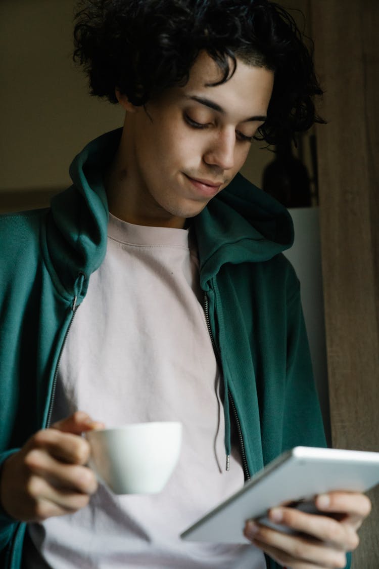 Young Ethnic Man Reading Tablet And Drinking Coffee Near Window At Home