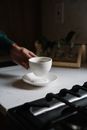 Crop unrecognizable person holding cup of coffee above table with plate in dark kitchen