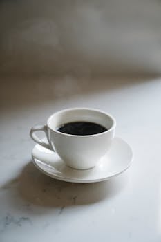 A hot cup of coffee in a white ceramic cup with steam rising, placed on a marble countertop.