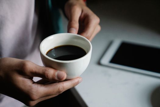 Close-up of a person holding a cup of coffee near a tablet on a table.