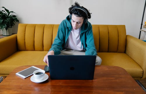 Free High angle of busy young male in headphones sitting on sofa and using cellphone at table with netbook coffee and tablet Stock Photo