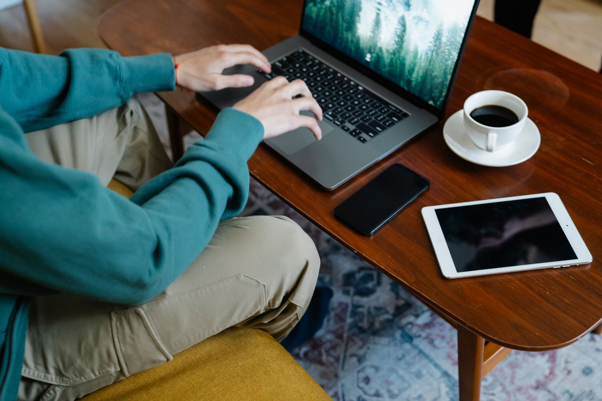 High angle of crop anonymous male remote worker working on netbook at table with cup of coffee placed near cellphone and tablet