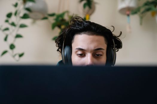 A young man wearing headphones intensely focused on work at a computer indoors with plants in the background.