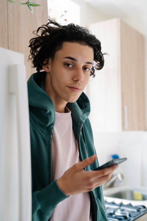 Serious young curly haired male in casual clothes standing near fridge and browsing cellphone while looking at camera