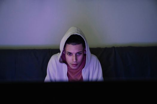 Focused young ethnic male in hood looking forward while sitting on couch in house room