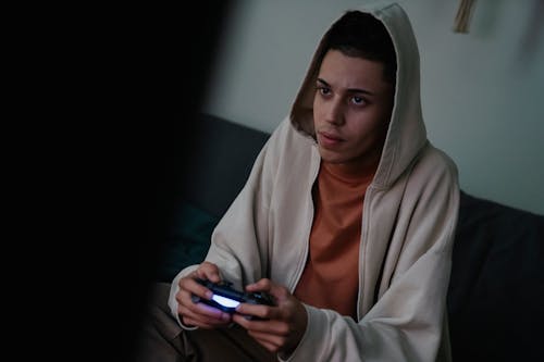 Addictive young ethnic male gamer in hoodie with console controller playing video game while looking forward at home
