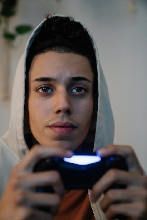 Concentrated young ethnic man in hood with console controller playing video game while looking forward in house room
