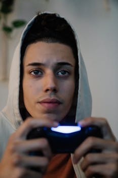Concentrated young ethnic man in hood with console controller playing video game while looking forward in house room