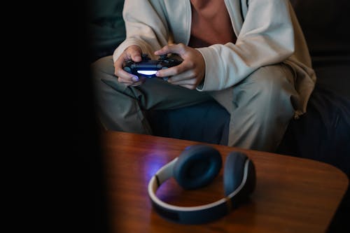 Crop unrecognizable man with joystick playing video game while sitting against table with modern headset in house room