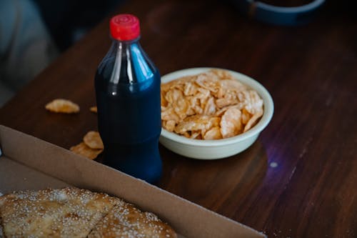From above of plastic bottle of soft drink between takeaway pizza with sesame seeds and crispy potato chips at home