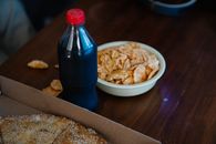 From above of plastic bottle of soft drink between takeaway pizza with sesame seeds and crispy potato chips at home