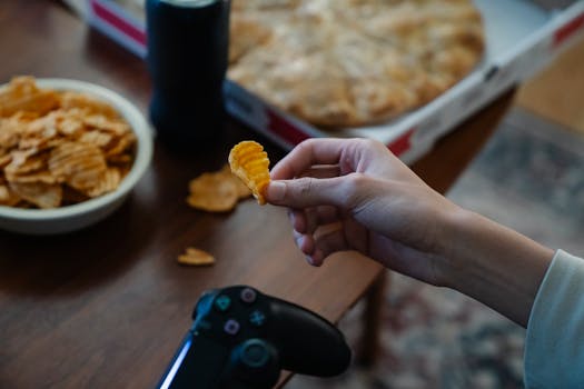 A person holding a chip while playing a video game with snacks and pizza nearby.