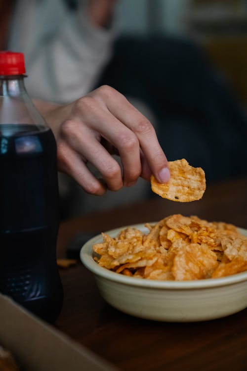 Free Crop anonymous person against table with crunchy potato chips and bottle of soda in house room Stock Photo
