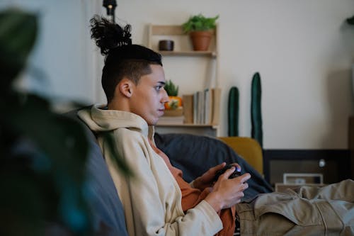 Side view of ethnic male gamer with hair bun and console controller playing video game on sofa in living room