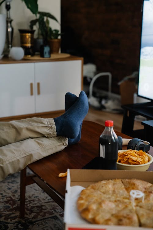 Crop anonymous person with crossed legs on table against soft drink and potato chips in house