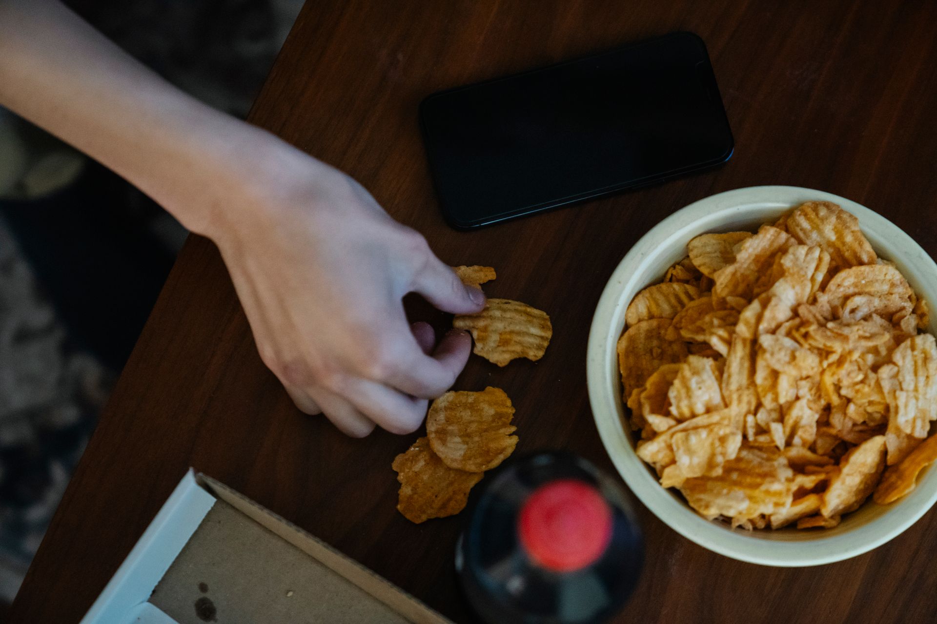 From above of crop anonymous person with crispy potato chips at table against bottle of refreshing drink and smartphone