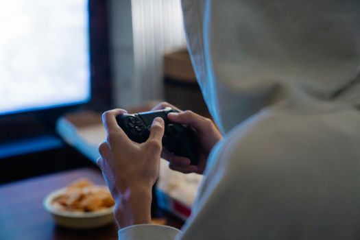 Back view of crop anonymous man in hoodie pressing button on joystick while playing video game against monitor