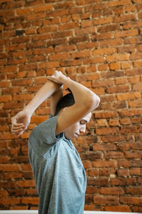 Side view of male teenager in t shirt stretching out arms and closing eyes against brick wall