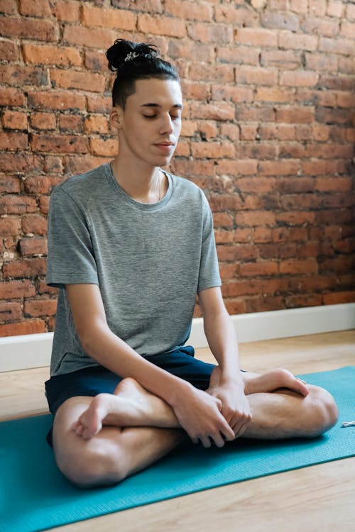 Full body of calm teenager in sportswear sitting on mat while doing Padmasana against brick wall