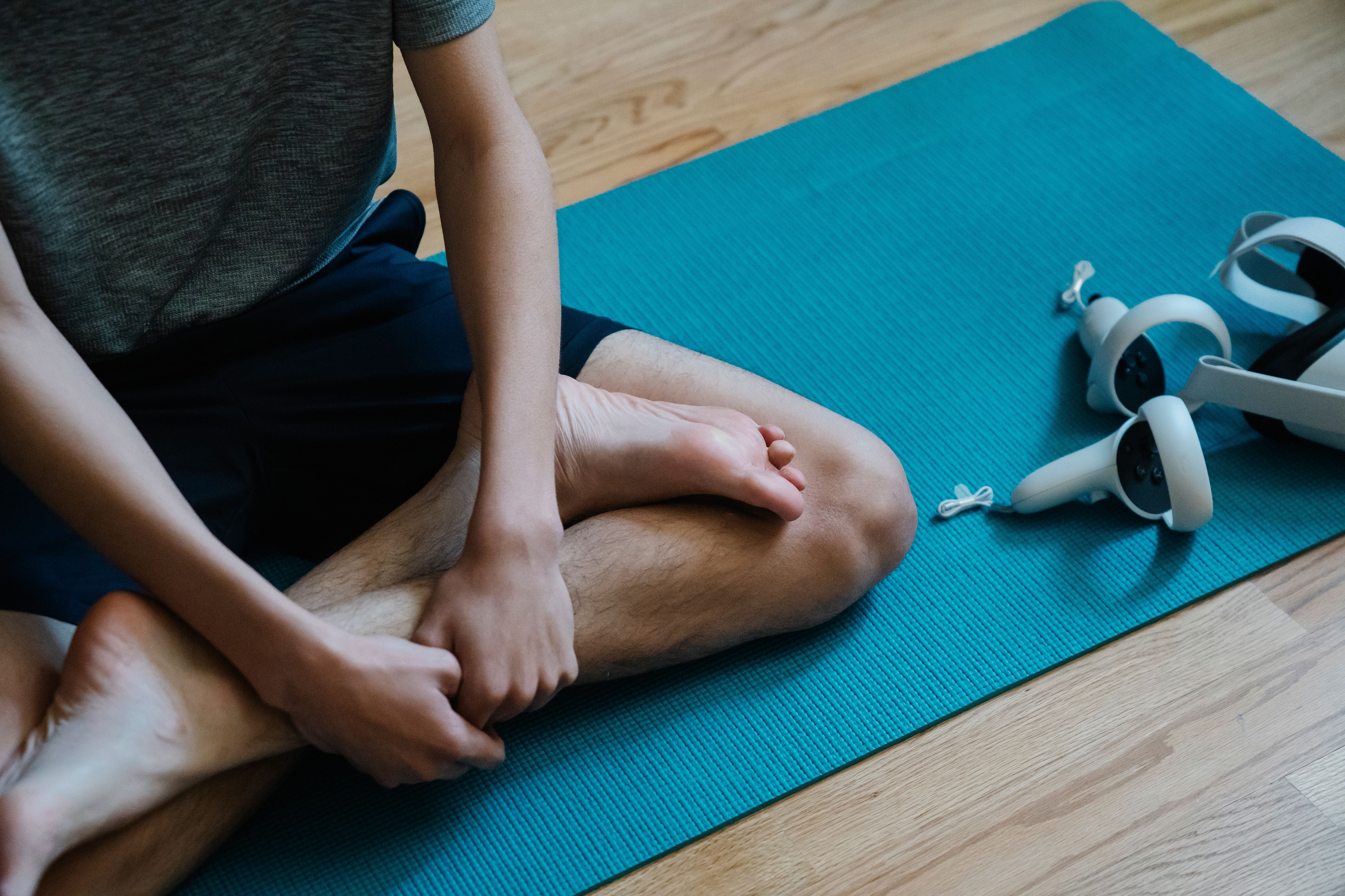 A person sits cross-legged on a turquoise yoga mat with a VR headset and controllers resting nearby on a wooden floor.
