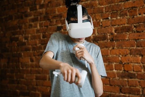 Free Young male teenager in casual clothes using VR headset and controllers while playing game near brick wall Stock Photo