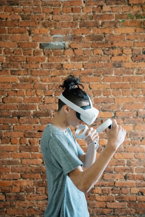 Side view of young male gamer in casual outfit using VR headset and controllers in light room with brick wall