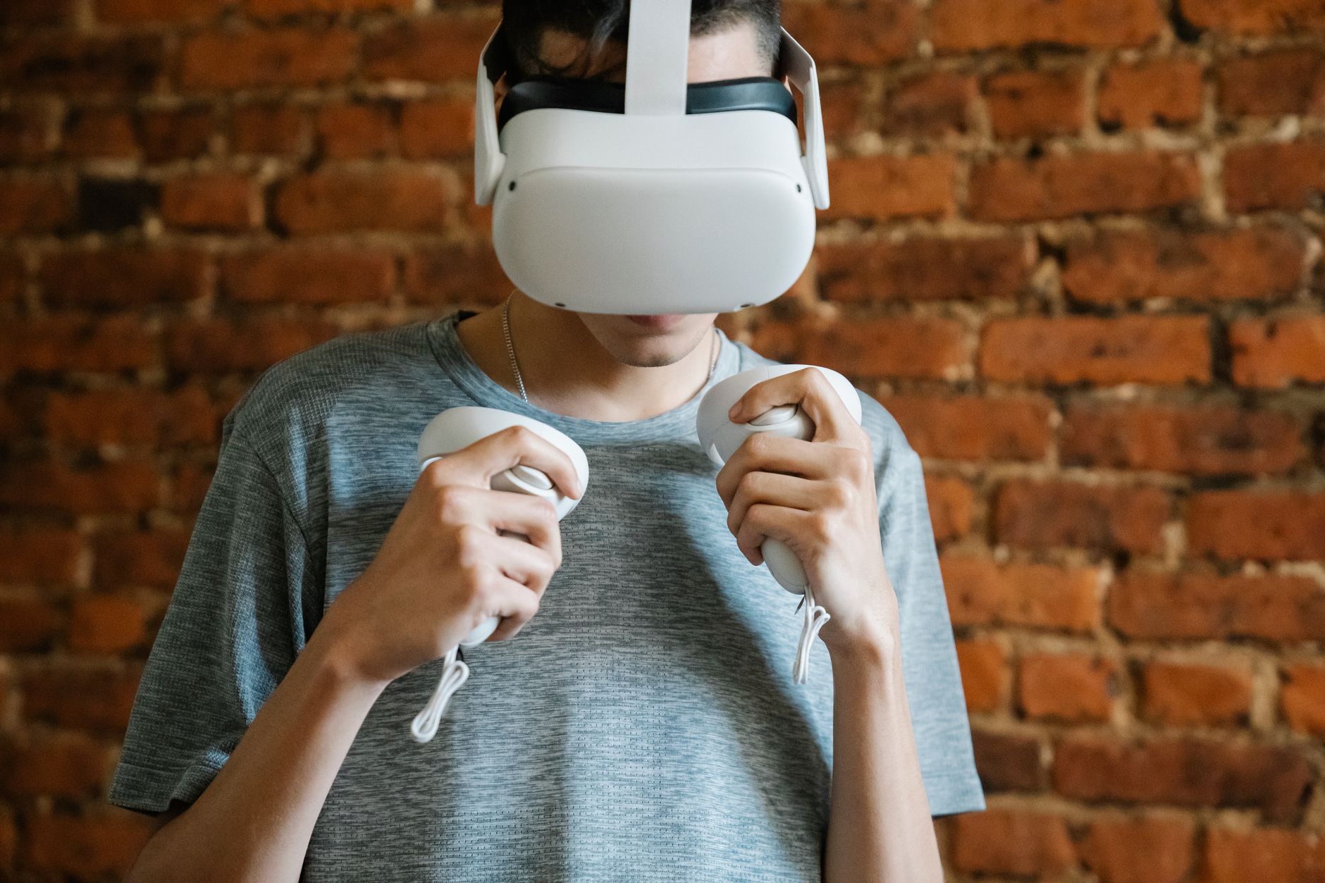 Young unrecognizable male in gray casual t shirt experiencing virtual reality with headset and controllers on background of brick wall