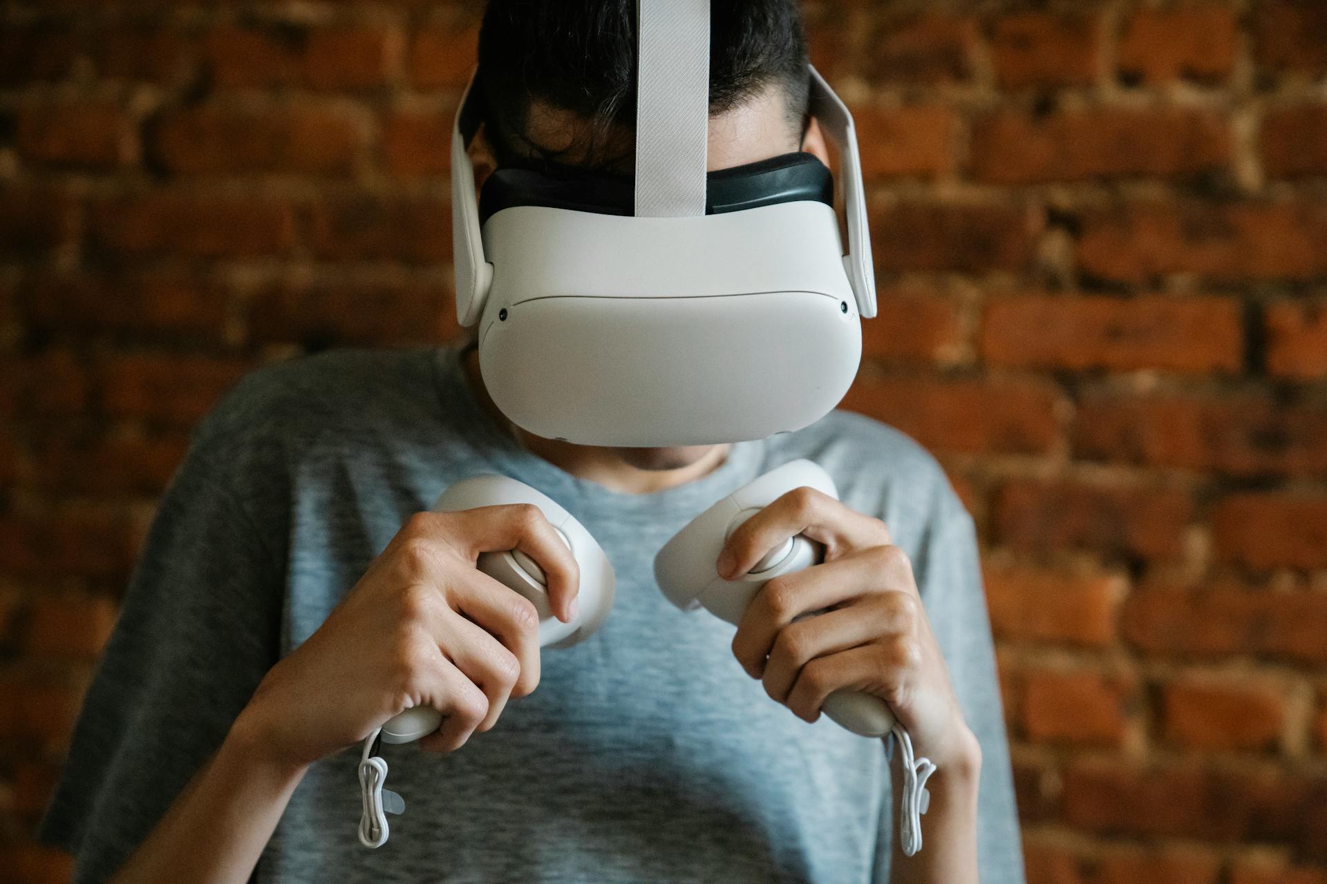 Crop anonymous male in casual clothes and virtual reality goggles with controllers playing video game against brick wall